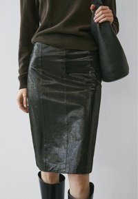 Jupe crayon en cuir noir avec une surface brillante et texturée, présentant des coutures verticales et une silhouette cintrée, associée à un sac noir texturé.