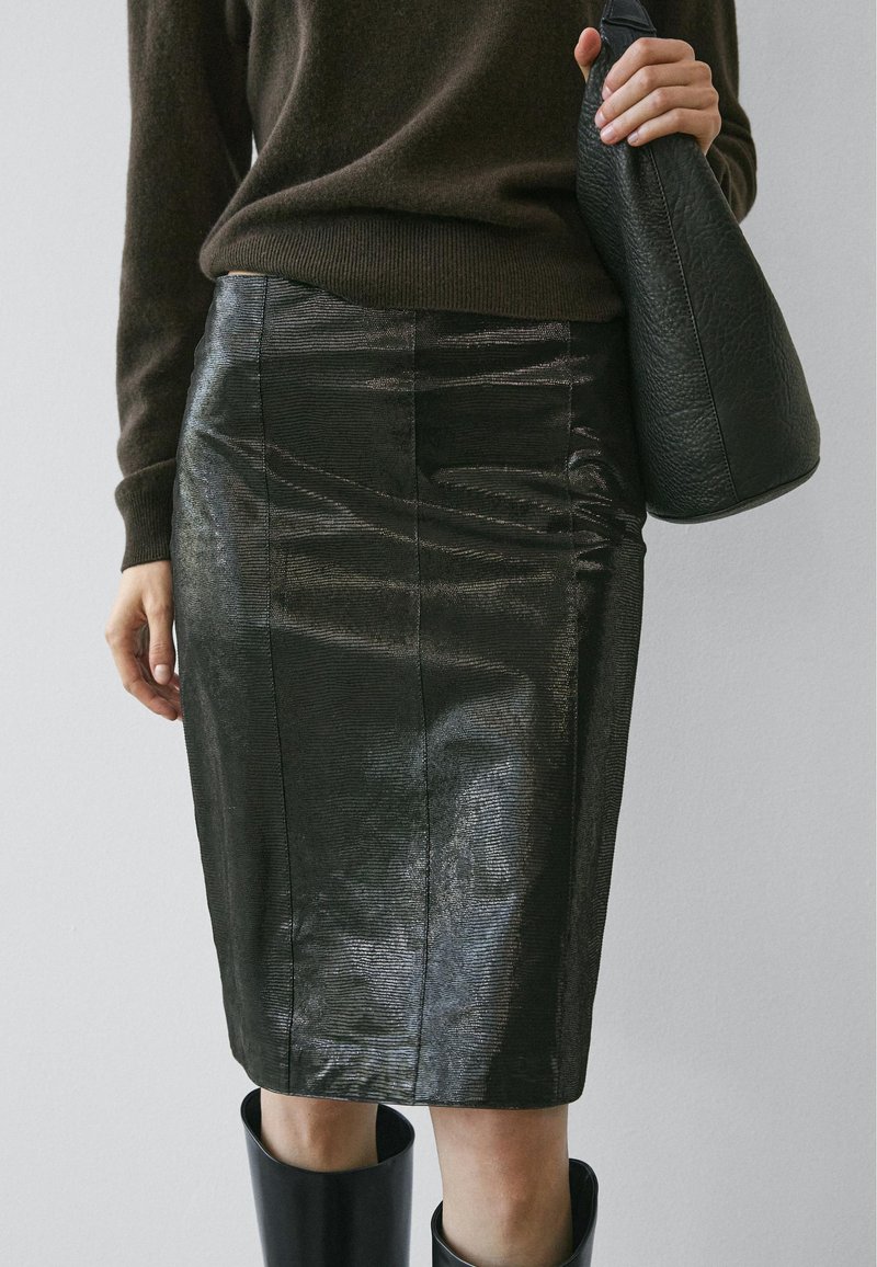 Jupe crayon en cuir noir avec une surface brillante et texturée, présentant des coutures verticales et une silhouette cintrée, associée à un sac noir texturé.