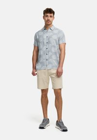 Camisa de manga corta de color azul claro con un patrón de hojas blancas, con botones en el frente, combinada con pantalones cortos beige y zapatillas grises.