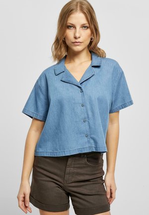 Lichtblauwe spijkerhemel met korte mouwen, een kraag, knoopjes aan de voorkant en een cropped design. Gecombineerd met donkerbruine shorts.