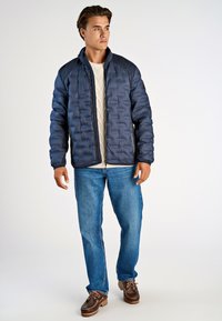 Marinblå pufferjacka med quiltat mönster, ståkrage och dragkedja, bärs över en krämfärgad kabelstickad tröja och blåjeans.