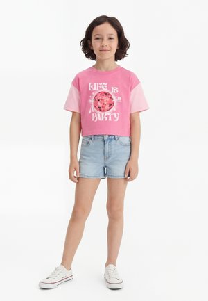 Barn iført en rosa cropped T-skjorte med teksten "Life is a party" og diskokule-grafikk, lyse denimshorts og hvite joggesko, stående.
