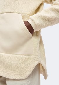 Main à la peau foncée dans la poche d'une veste polaire crème avec un ourlet incurvé et des poignets côtelés.
