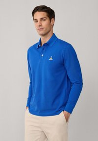 Langärmliges Polohemd in leuchtendem Blau, aus weicher Baumwolle gefertigt. Verfügt über einen Kragen, eine Drei-Knopf-Leiste und ein kleines gesticktes Logo auf der Brust.