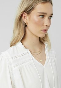 Blouse blanche texturée avec des accents en dentelle, en V et avec un cordon de serrage. Colliers en or superposés avec un pendentif et boucles d'oreilles pendantes avec un détail noir.