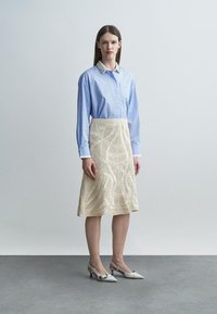 Γυναίκα που στέκεται φορώντας ένα μπλε ριγέ πουκάμισο με κολάρο από χάντρες, μια κρεμ υφασμάτινη φούστα μήκους έως τα γόνατα και άσπρα γοβάκια με τακούνι και δέσιμο πίσω.