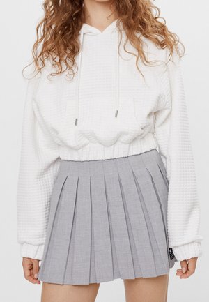 Kobieta w białej, teksturowanej bluzie z kapturem i wysokim stanem szarej plisowanej spódnicy na jednolitym tle.