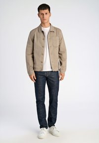 Beige bomullsjacka med framficka, knäppning framtill, bärs över en vit T-shirt, i kombination med mörkblå jeans och vita sneakers.