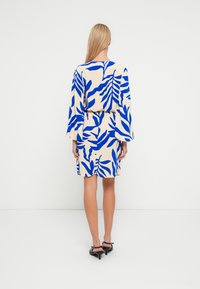 Femme aux longs cheveux blonds portant une robe blanche avec des motifs de feuilles bleues et des sandales à talons noires, vue de dos sur un fond blanc uni.