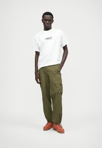 Biela tričko s krátkymi rukávmi a čiernym logom, olivovo zelené cargo nohavice s bočnými vreckami a oranžové šnurovacie topánky s textúrovanou podrážkou.