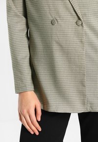 Dwurzędowa marynarka w kratę w odcieniach jasnobeżowym i czarnym, z długimi rękawami i teksturowaną tkaniną.