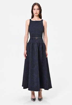 Donna in piedi che indossa un abito midi blu navy senza maniche, in tessuto strutturato, con cintura in vita e décolleté grigie con punta.