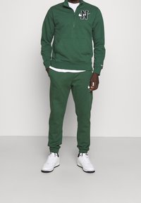 Grön zip-up sweatshirt med svart logotyp på bröstet och fickor, matchad med joggers i samma färg. Bärs med vita sneakers. Slät textur.