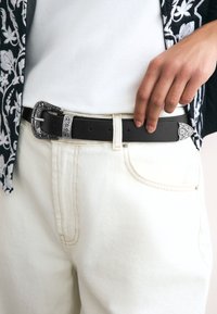 Cintura in pelle nera con fibbia decorativa in argento, caratterizzata da incisioni floreali e una punta appuntita, indossata sopra pantaloni di denim bianchi.