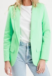 Blazer vert menthe à coupe ajustée, avec deux poches avant et une texture lisse, porté avec un t-shirt blanc et un jean bleu clair.