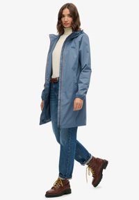 Niebieski wodoodporny płaszcz przeciwdeszczowy z kapturem, z zamkiem z przodu i elastycznymi mankietami. W połączeniu z jasnoniebieskimi dżinsami i brązowymi butami turystycznymi.