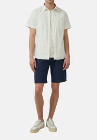 Camicia leggera a maniche corte con motivo a foglia, pantaloni corti blu navy e sneakers bianche con dettagli beige. Materiale in cotone, vestibilità rilassata, design informale.