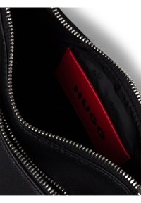Bolso de cuero negro con cremallera plateada. El interior muestra un tarjetero rojo con la palabra "HUGO" en negro. Superficie texturizada, diseño minimalista.