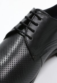 Chaussure habillée en cuir noir avec un bout perforé et un système de laçage, sur fond blanc.