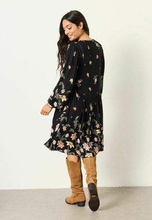 Frau lächelt und geht weg, trägt ein schwarzes Kleid mit Blumenmuster und langen Ärmeln sowie hellbraune kniehohe Wildlederstiefel.