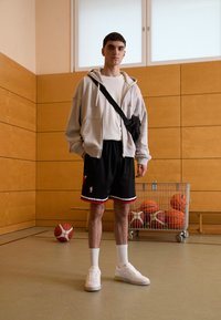 Completo sportivo composto da una felpa grigia chiara, una maglietta bianca, pantaloni corti neri con dettagli rossi e scarpe bianche, ambientato in una palestra con palloni da basket.