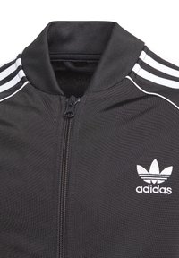 Černá bunda na zip z hladké látky. Má vysoký límec a tři bílé pruhy na ramenou, s bílým logem Adidas.