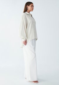 Camicia button-up beige chiaro con maniche lunghe e orlo curvo, abbinata a pantaloni bianchi a vita alta e ampia gamba, accompagnati da sandali minimalisti.