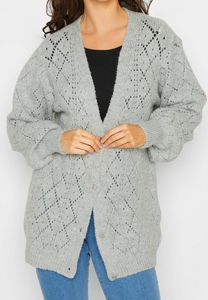 Kobieta zapinająca guziki szarego, dzianinowego kardiganu z rombowym wzorem, ubrana w czarny top i niebieskie dżinsy.