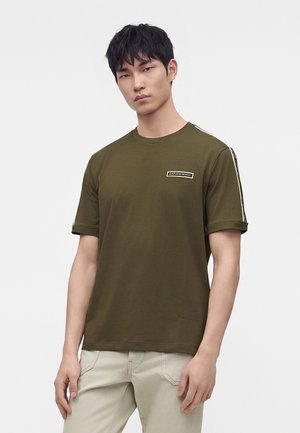 Homme portant un t-shirt vert olive à manches courtes avec un logo sur la poitrine et des rayures sur les manches, associé à un pantalon beige clair, debout.