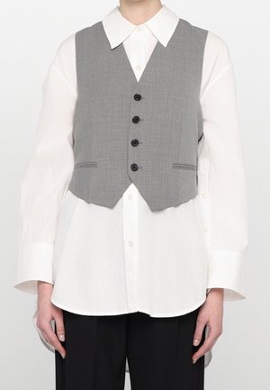 Osoba ubrana w białą, zapinaną na guziki koszulę z długim rękawem i szerokimi mankietami oraz szary, zapinany na guziki kamizelkę, do tego czarne spodnie.