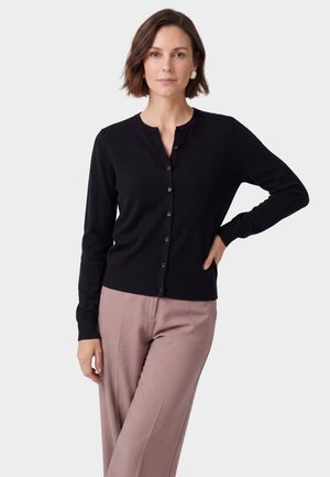 Femme aux cheveux courts bruns porte un cardigan noir boutonné et un pantalon marron clair, debout avec une main sur la hanche devant un fond uni.