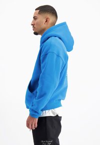 Blauwe hoodie met een losse pasvorm, voorzien van een buikzak en ribgebreide manchetten. Draagt boven een wit shirt en gecombineerd met donkere broek.