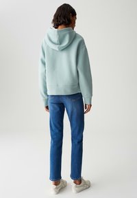 Persona di spalle che indossa un felpa azzurro chiaro, jeans blu e sneakers bianche, appoggiata su uno sfondo chiaro e neutro.