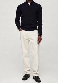 Marineblauer Rippstrickpullover mit einem Viertel-Reißverschluss-Kragen, kombiniert mit weißen geraden Hosen und schwarzen Lederschuhen.