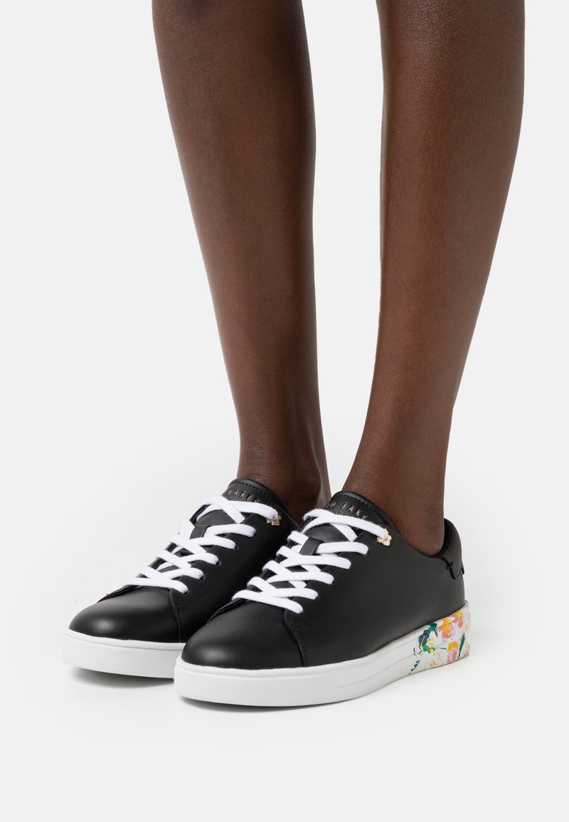 Schwarze Leder-Sneaker mit weißen Schnürsenkeln, die ein florales Muster auf der Sohle aufweisen. Abgerundete Zehenform und minimalistisches Design.