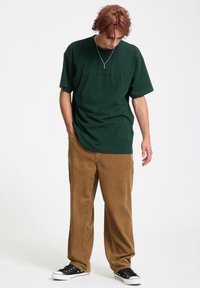 Zielona, oversize'owa bawełniana koszulka z logo "Volcom", zestawiona z dopasowanymi, brązowymi spodniami z korduroju. Czarne kanwasowe trampki dopełniają strój.
