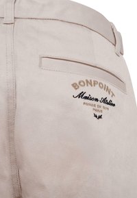 Pantaloni in cotone beige con una tasca posteriore che presenta un ricamo con la scritta "BONPOINT Maison Atelier" in filo dorato e nero. Texture liscia.