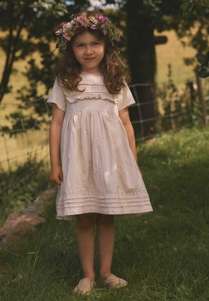 Nuori kiharatukkainen tyttö valkoisessa mekossa ja kukkaseppele päässään seisoo ruohikolla metalliaidan lähellä varjossa ulkona.