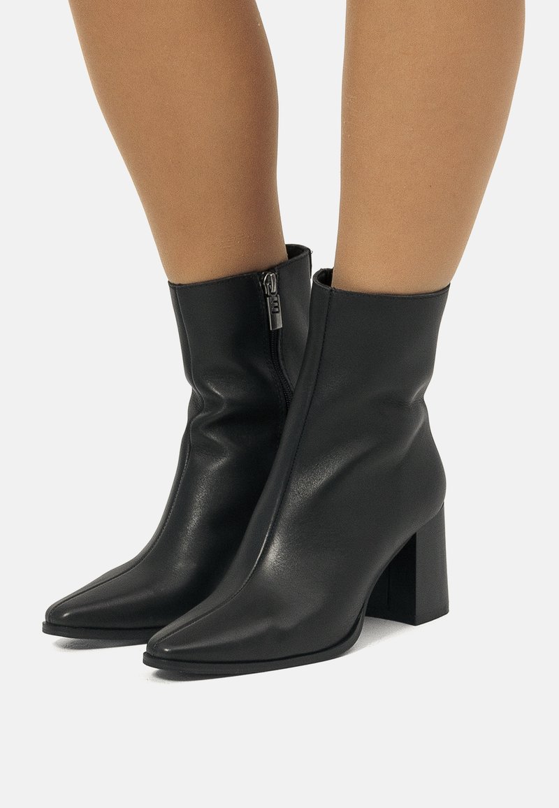 Schwarze Leder-Ankle-Boots mit seitlichem Reißverschluss, spitzer Zehenform und Blockabsätzen, getragen auf nackten Beinen vor einem schlichten Hintergrund.