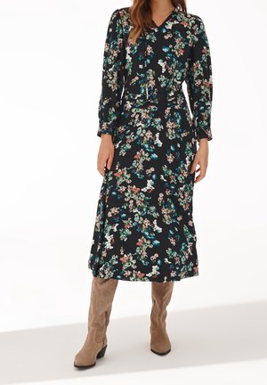 Vrouw draagt een zwarte midi-jurk met lange mouwen en bloemenprint, met een gestrikte halslijn, gecombineerd met beige suède kniehoge laarzen.