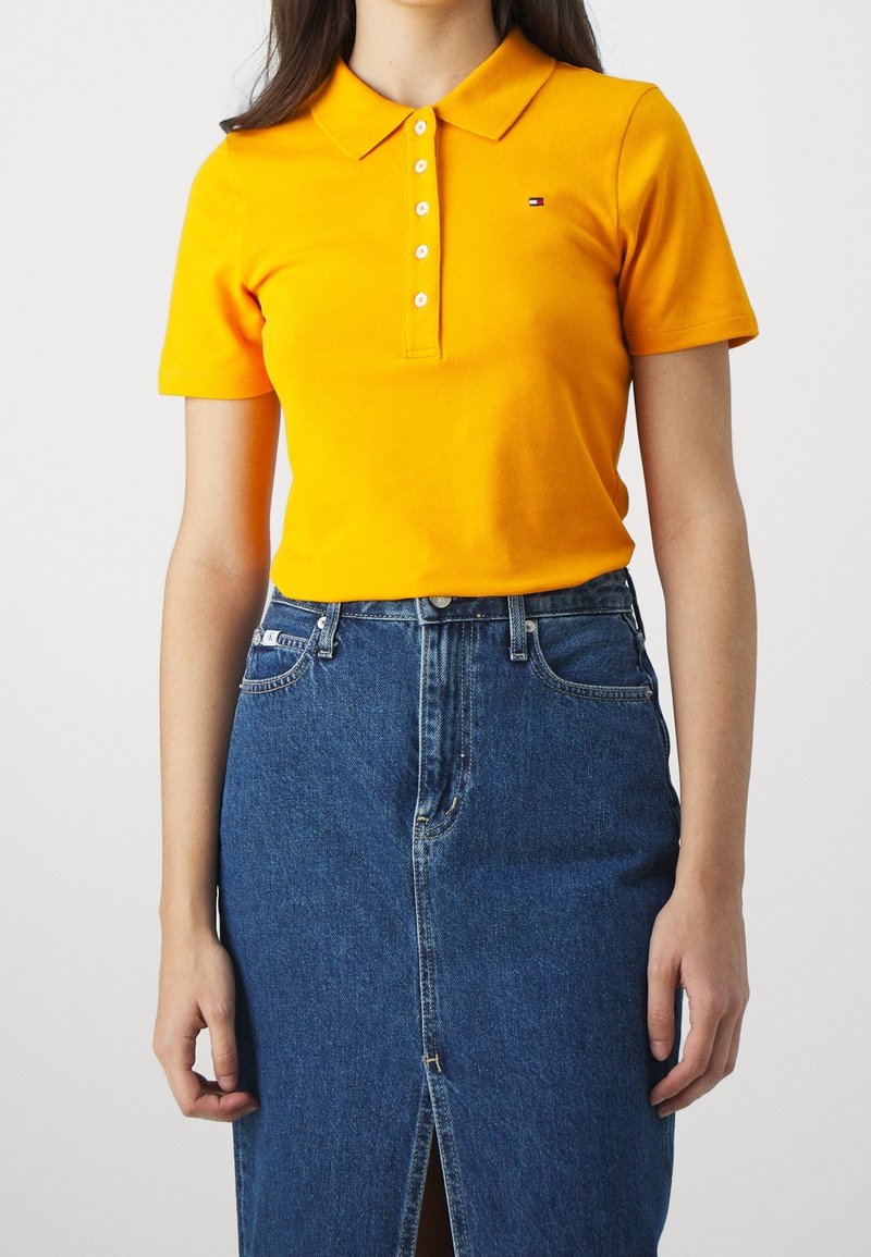 Femme portant un polo jaune vif à manches courtes avec des boutons et une jupe en denim bleu avec une fente à l'avant, debout devant un fond uni.