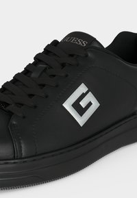 Baskets en cuir noir avec lacets noirs et logo argenté "G" sur le côté, avec le nom de la marque "GUESS" sur la languette.