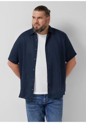 Bärtiger Mann mit zu einem Dutt gebundenen Haaren, trägt ein marineblaues kurzärmeliges Hemd über einem weißen T-Shirt und blaucblauen Jeans, steht mit den Händen hinter dem Rücken.