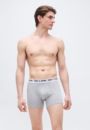 Νεαρός άνδρας με κοντά μαλλιά στέκεται χωρίς μπλούζα φορώντας γκρι μπόξερ με τη ζώνη μέσης «ONLY & SONS», κοιτάζοντας προς τα δεξιά, σε απλό φόντο.