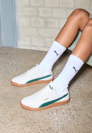 Beine in weißen Puma-Sneakern mit grünen Streifen und braunen Sohlen, kombiniert mit weißen Socken mit Puma-Logo, sitzend auf einem strukturierten Boden.