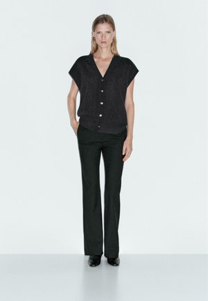 Black short-sleeve top with button closure and a relaxed fit, paired with fitted dark green trousers and black shoes, set against a plain background.