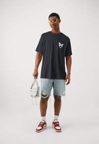 Black cotton t-shirt with a white butterfly graphic on the left chest, paired with light blue denim shorts, white and red sneakers, and a small gray bag.