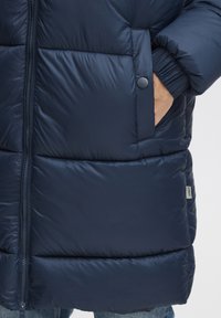 Giacca puffer blu navy con chiusura a zip frontale, pannelli a costine orizzontali, polsini elasticizzati e tasca laterale con chiusura a scatto. Materiale testurizzato.