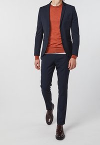 Marinblå kostym med skräddarsydd passform, åtföljd av en rostfärgad tröja. Bruna läderskor med slät finish fulländar looken.