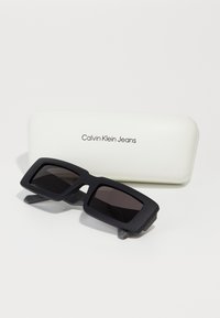 Ochelari de soare dreptunghiulari negri cu lentile întunecate, având un finisaj mat și temple late. Împreună cu un toc alb inscripționat "Calvin Klein Jeans."